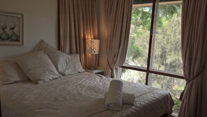 A neatly made bed with a rolled towel, a bedside lamp, and a view of trees through the window. - Linley House (Linley Point)