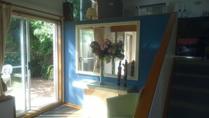 A room with a large sliding glass door leading to a garden, a blue wall with a window, and a small table with a vase of flowers. - Linley House (Linley Point)