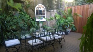 A glass-top table with black metal chairs and white cushions, surrounded by lush green plants and a wooden fence. - Linley House (Linley Point)