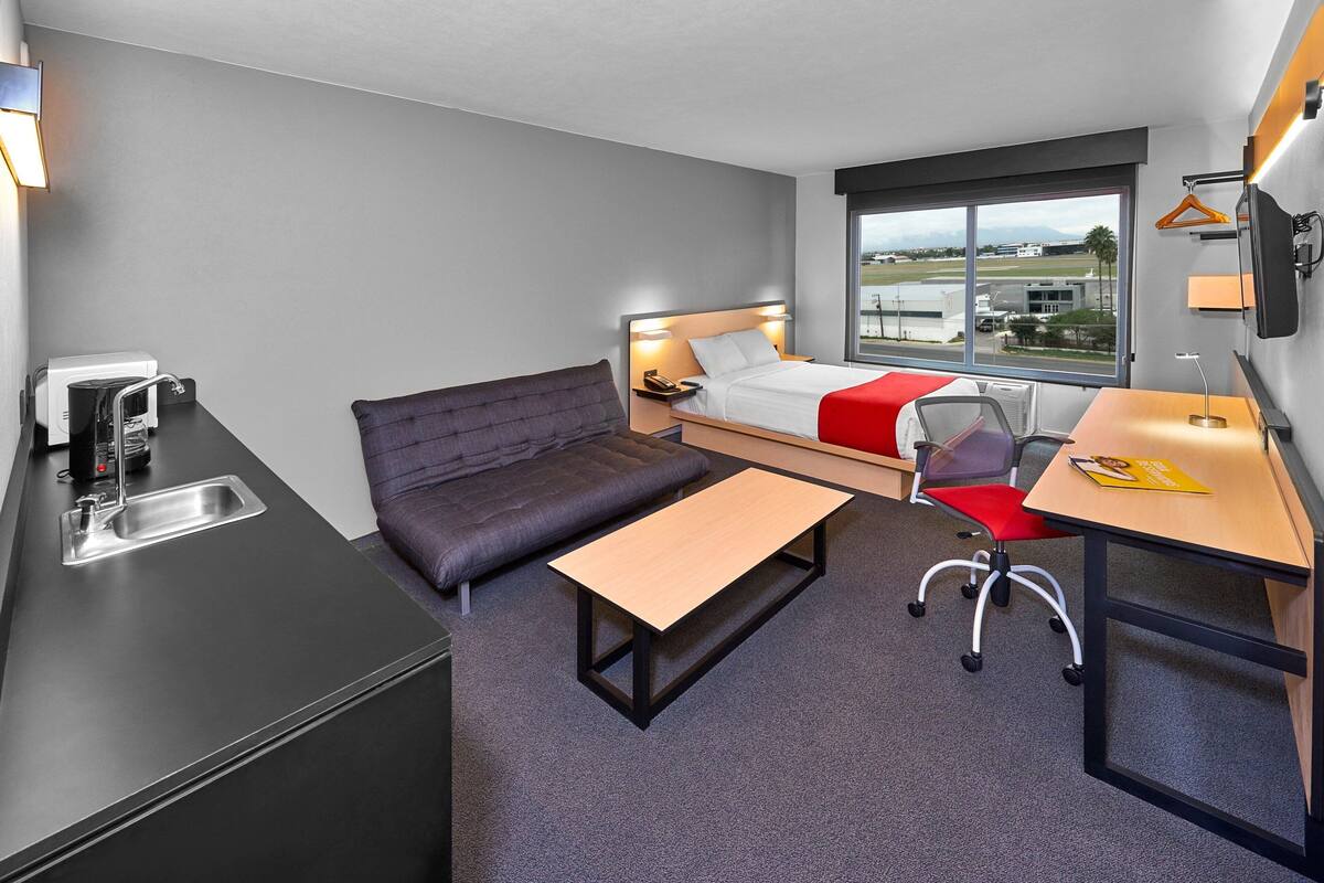 Habitación de hotel compacta con cama, sofá, escritorio y kitchenette.