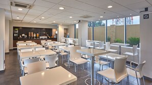Una cafetería amplia con mesas y sillas blancas, un mostrador con alimentos y grandes ventanales que ofrecen luz natural.