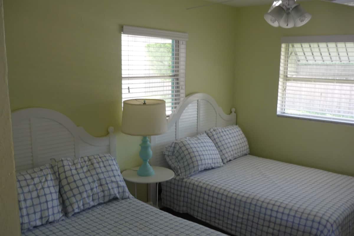 A room with two single beds, each with a headboard, a small round table with a lamp, and two windows with blinds.