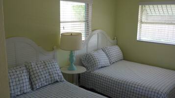A room with two single beds, each with a headboard, a small round table with a lamp, and two windows with blinds.
