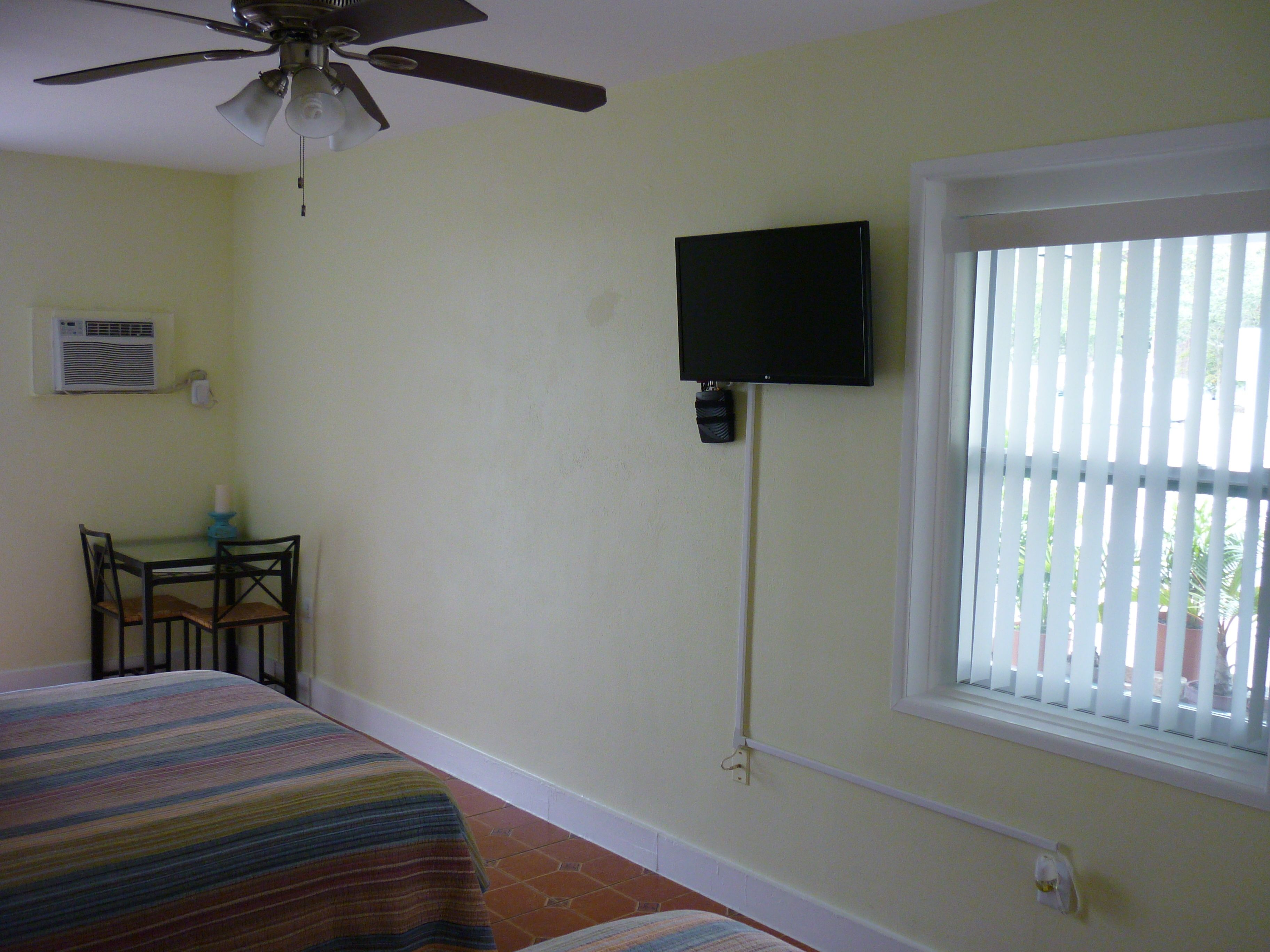 Une chambre avec un lit, un ventilateur de plafond, un téléviseur fixé au mur, une fenêtre avec des stores et une petite table avec des chaises.