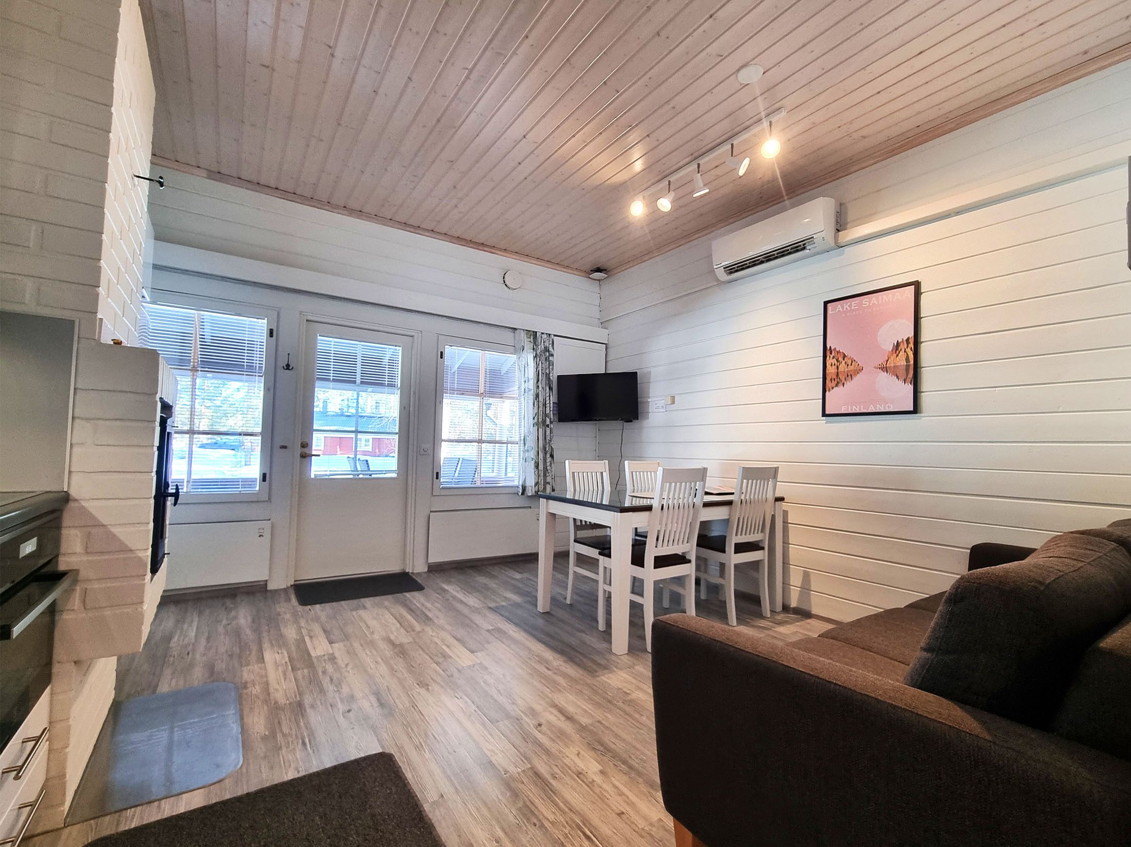 A room with a wooden ceiling, white walls, a dining table with chairs, a sofa, and a TV mounted on the wall.