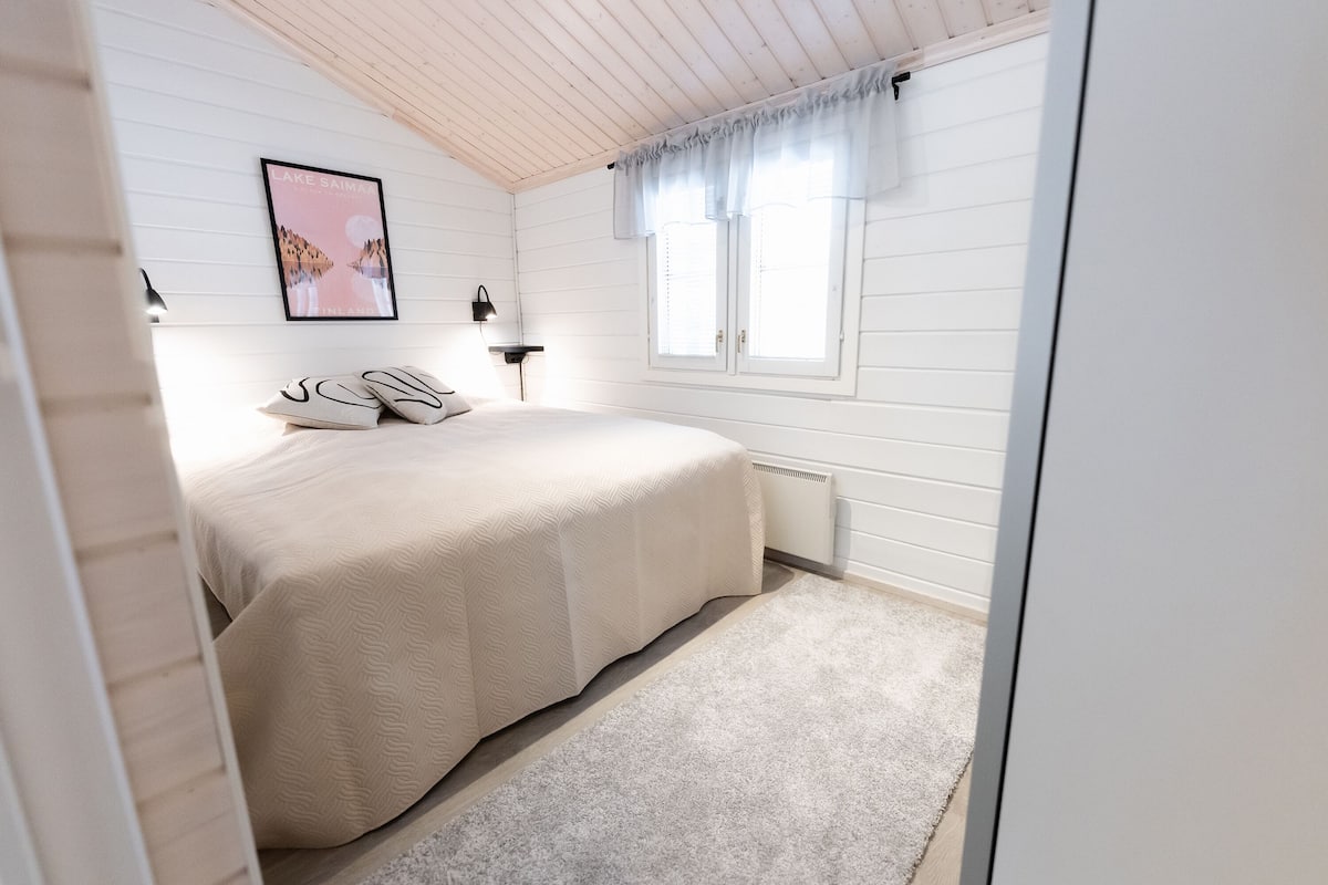 A bedroom with a bed, a bedside table, a lamp, a window with curtains, and a poster on the wall.