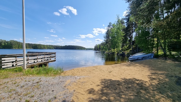 A lakeside view with a wooden dock, sandy beach, and a boat covered with a tarp. - Holiday Resort Harjun Portti (Savonlinna)
