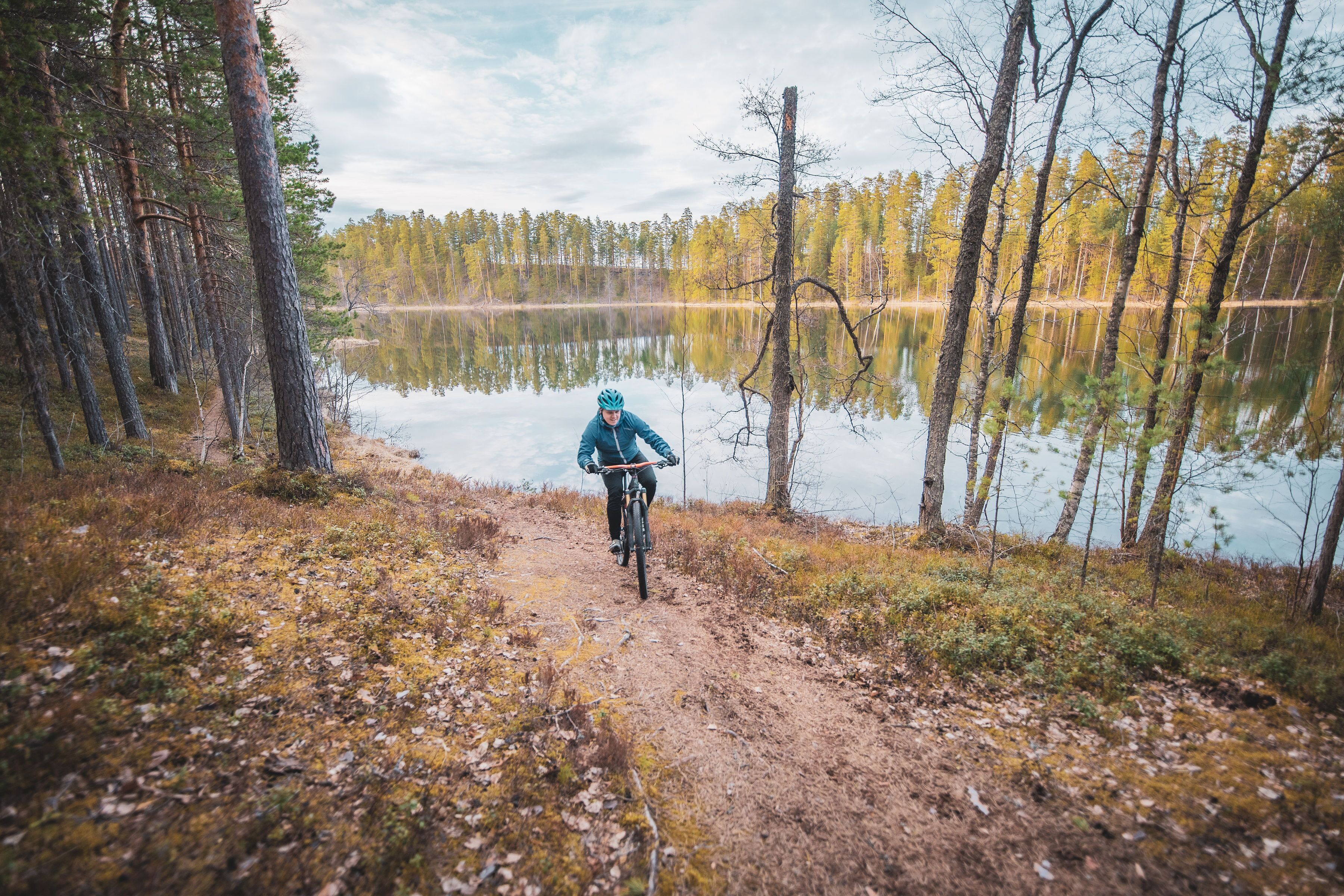 Un vététiste sur un sentier de terre, entouré d’arbres et bordé d’un lac.