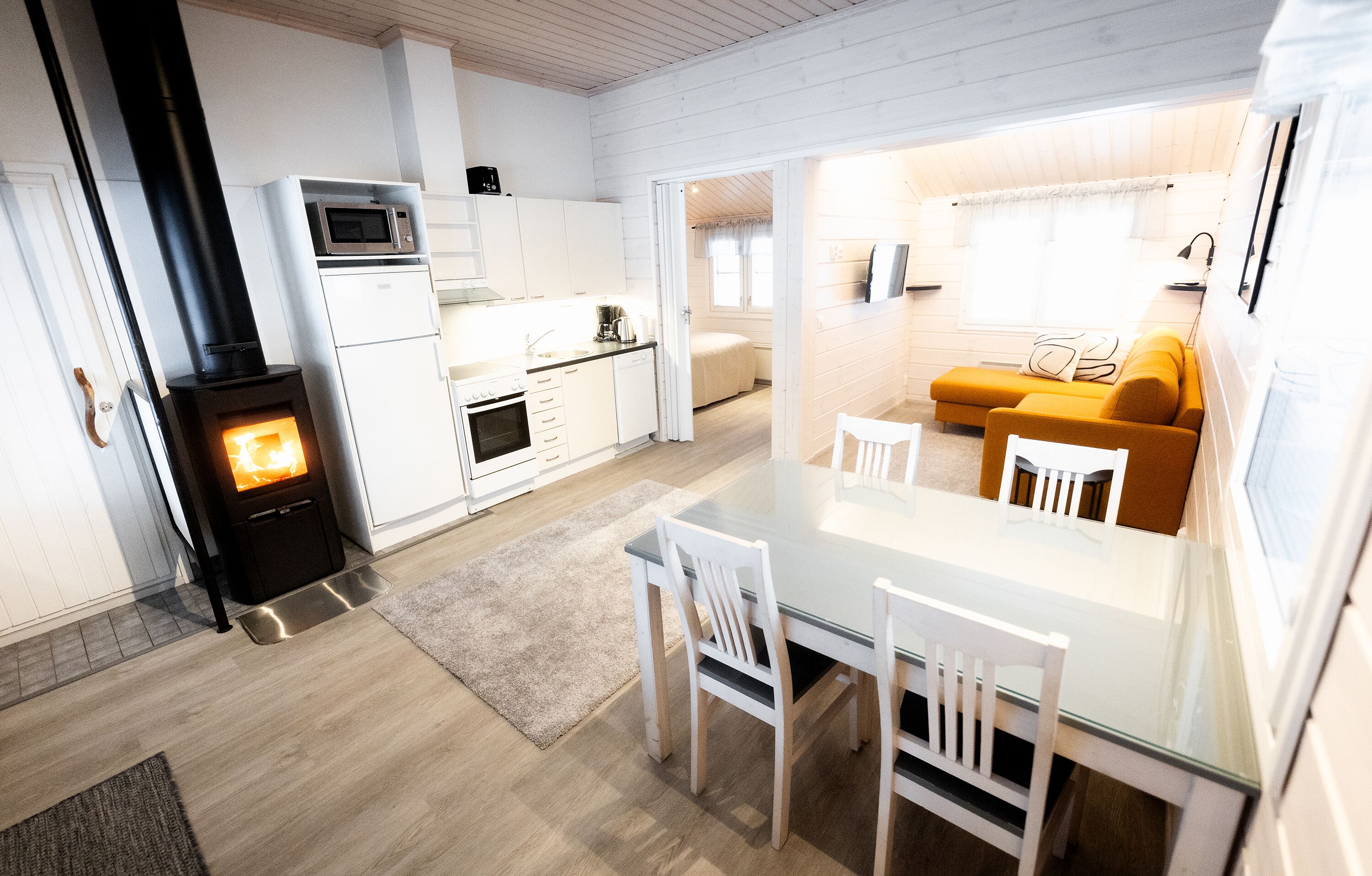 Een compacte, moderne keuken met een houtkachel, witte kasten en een eethoek met een glazen tafel en stoelen.