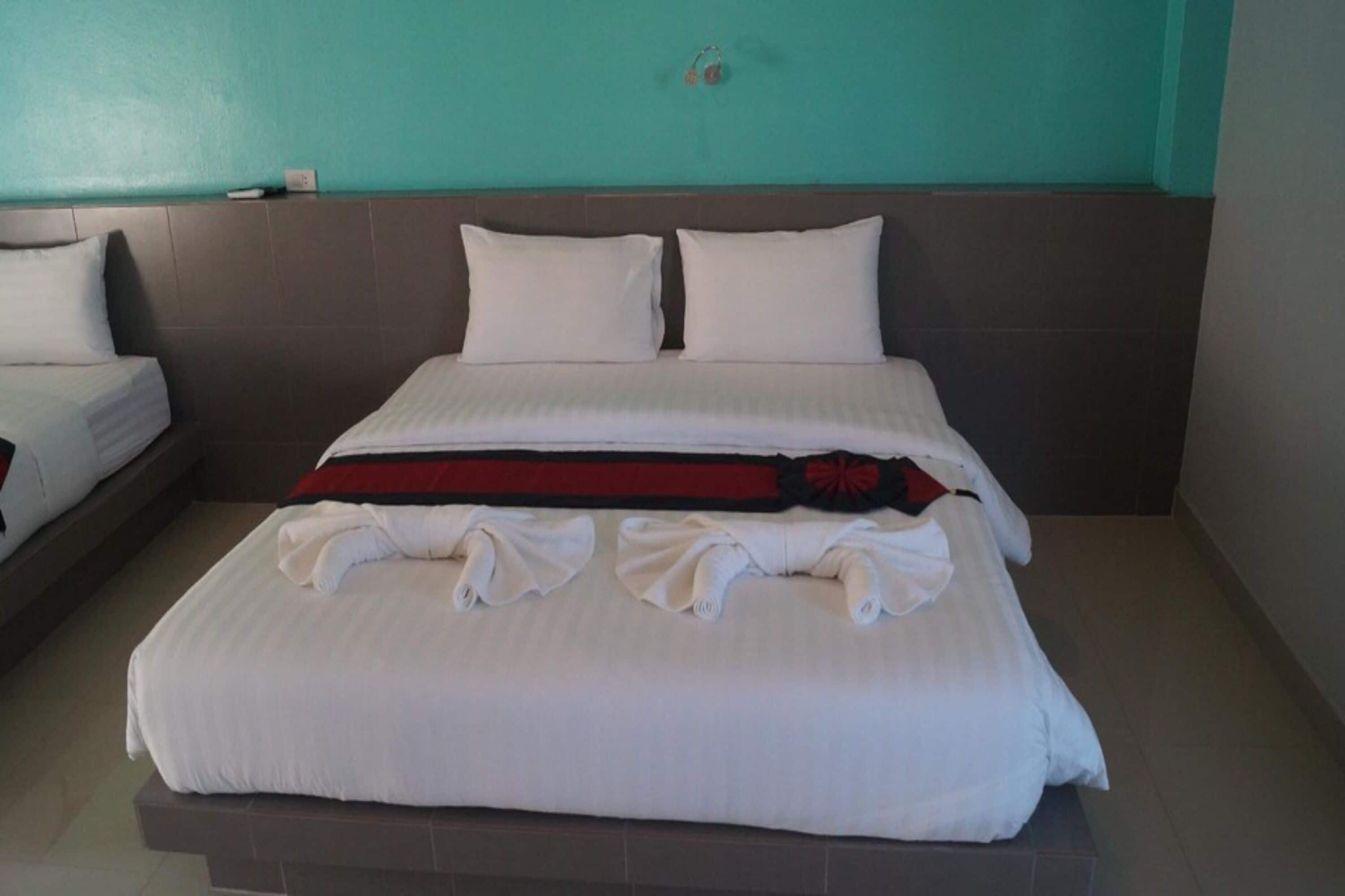 Habitación de hotel con una cama grande, dos almohadas, una manta roja y negra, y toallas dobladas con cuidado.