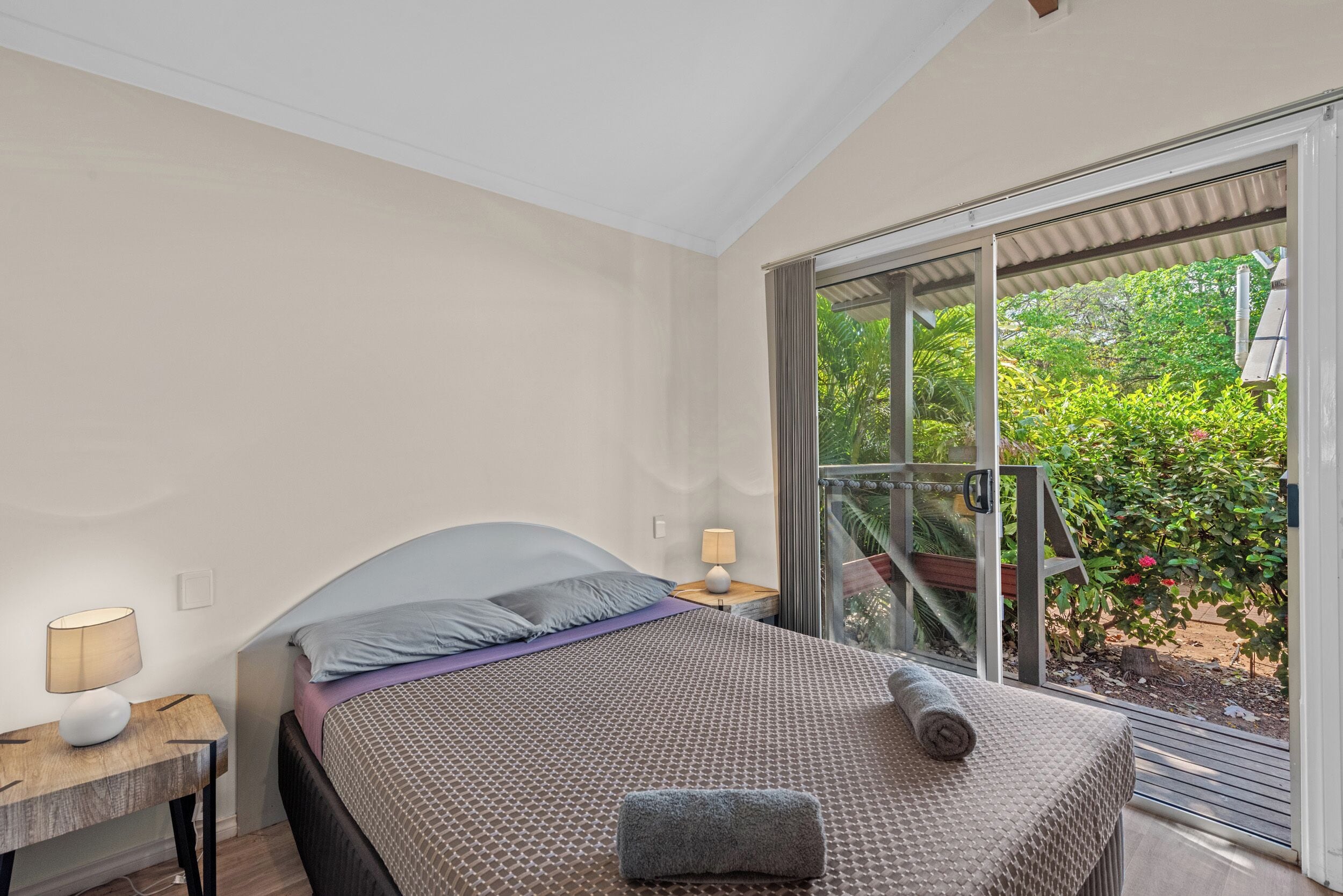 A bedroom with a bed, a nightstand, a lamp, and a view of greenery through a sliding door.
