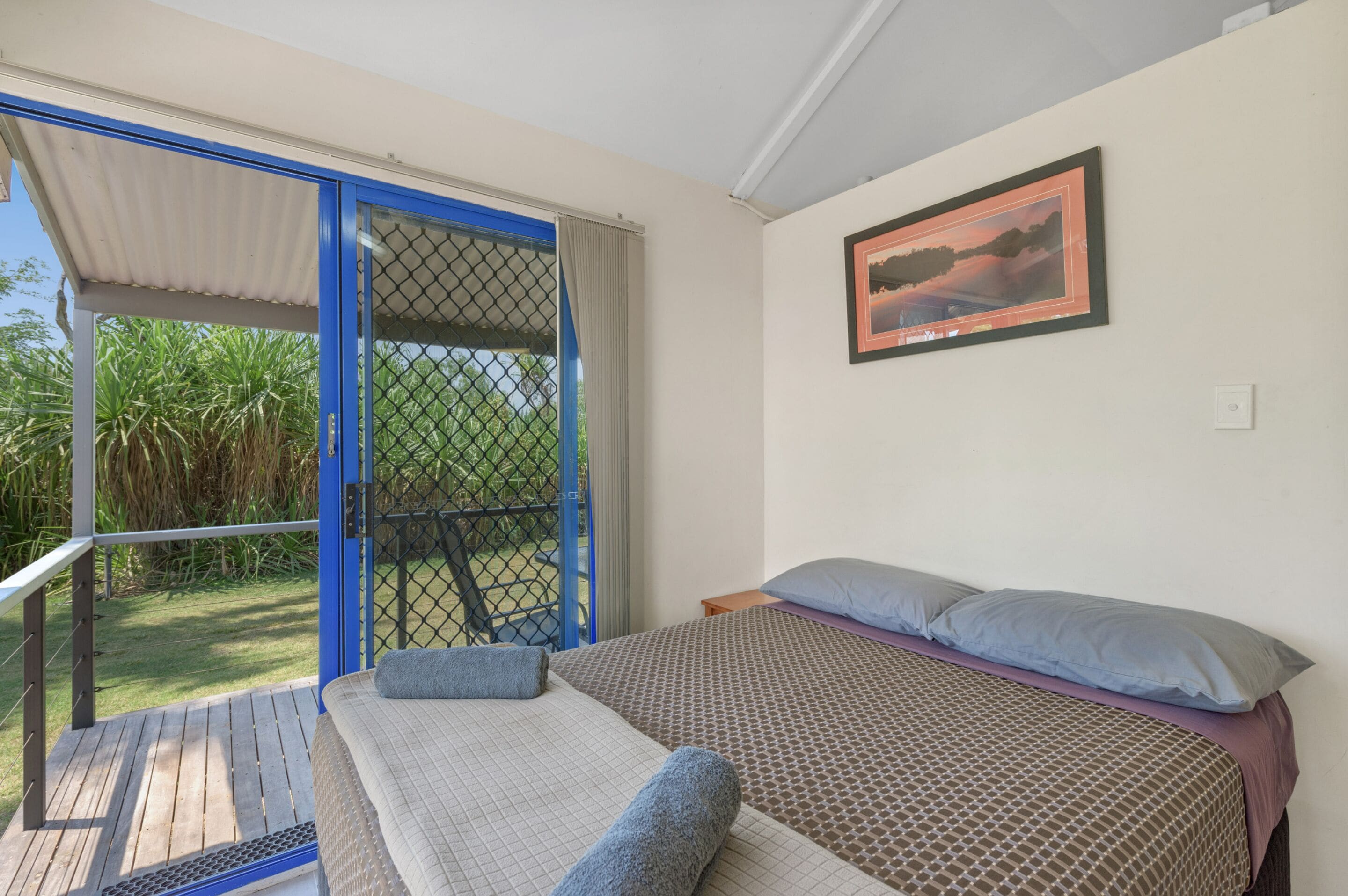 A bedroom with a bed, pillows, a framed picture on the wall, and a balcony with a view of greenery.