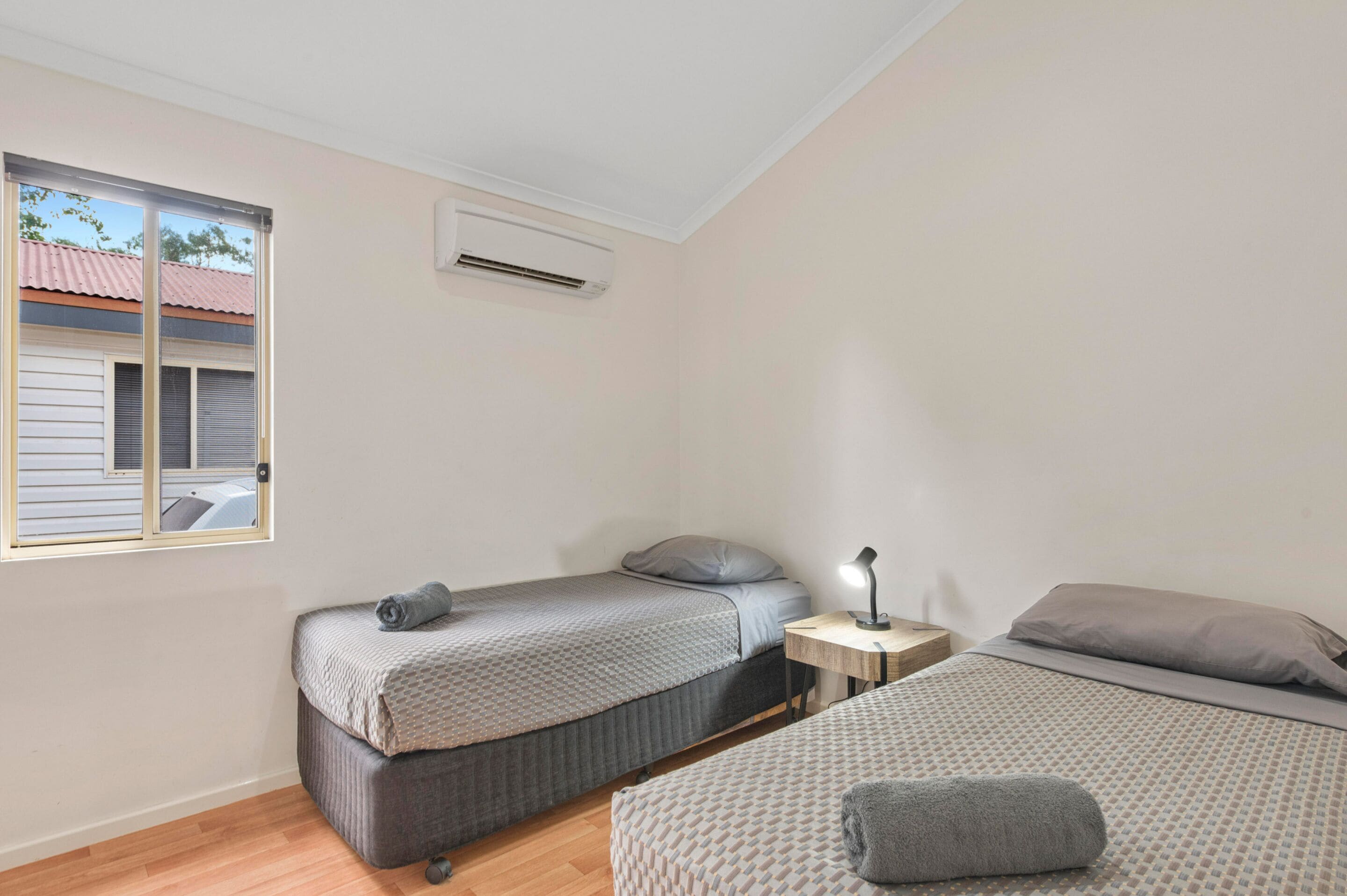 A small, clean room with two beds, a nightstand, and a window with blinds.