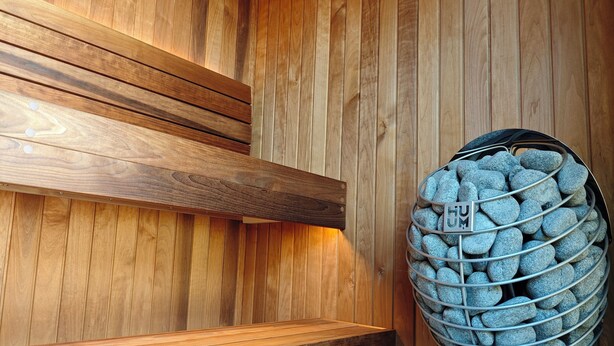 Un sauna en bois, avec un banc, un panier en métal rempli de pierres et un plancher en bois.