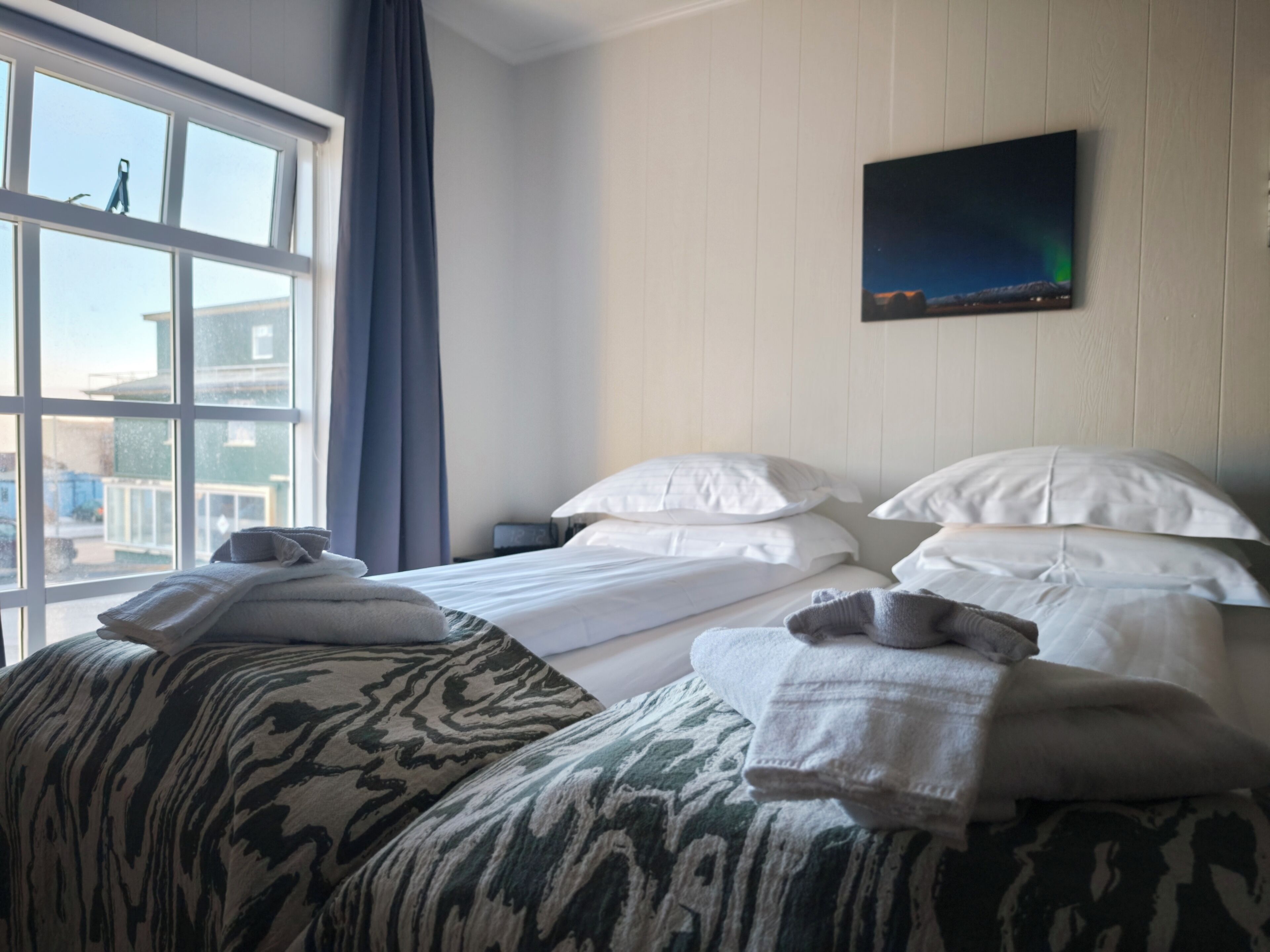 A hotel room with a bed, pillows, a blanket, a TV on the wall, and a window with curtains.