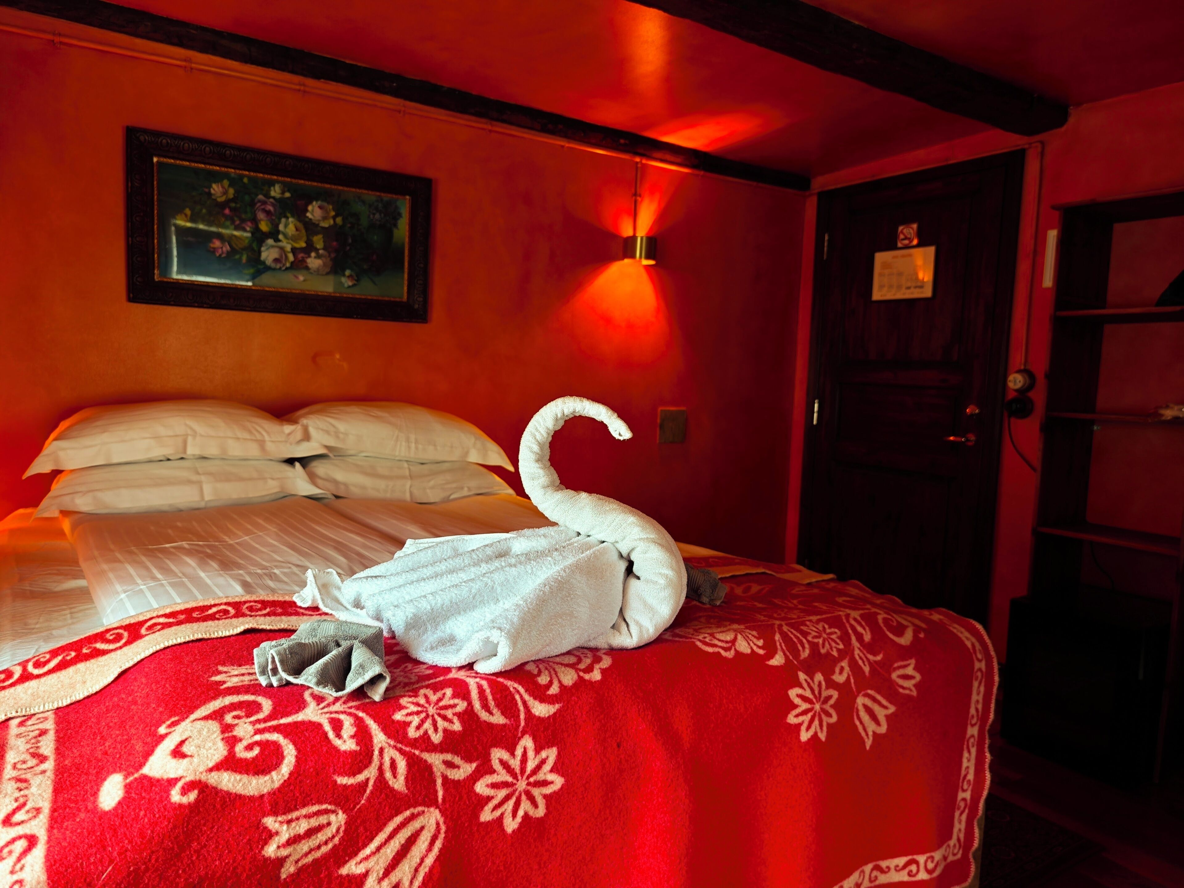 A hotel room with a bed, a red floral bedspread, a swan-shaped towel, a painting on the wall, and a door with a sign.