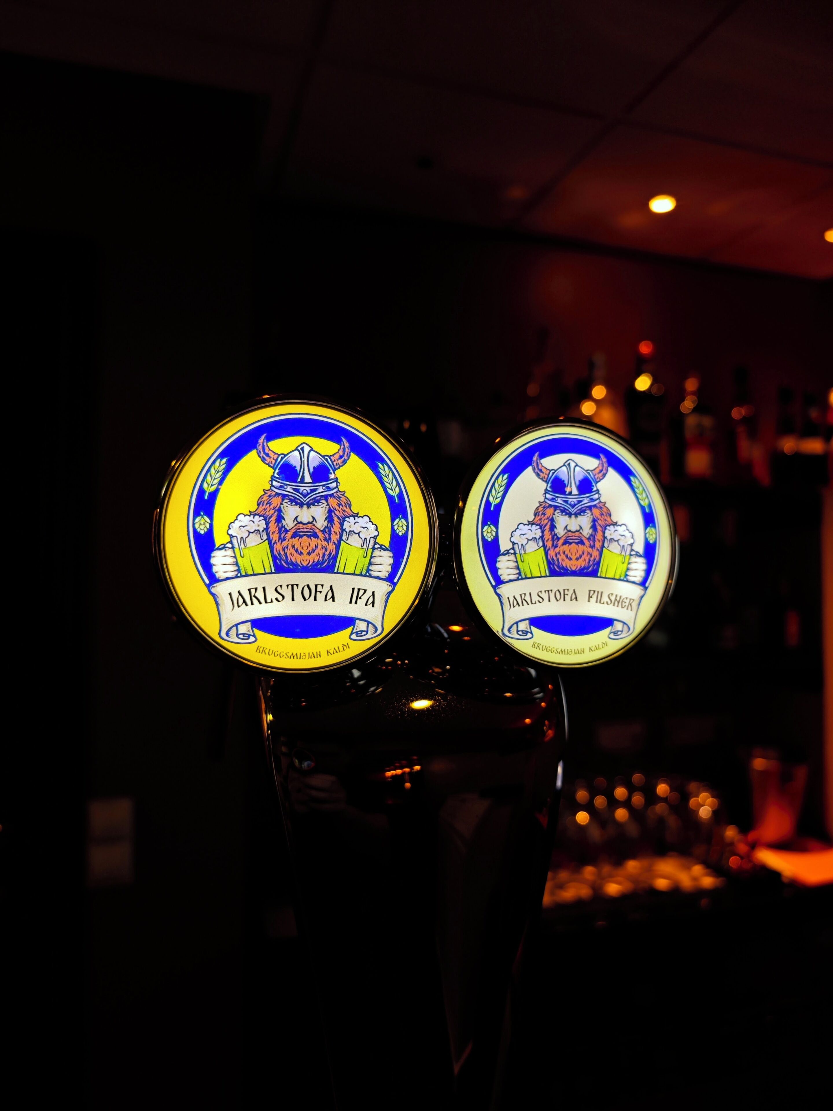 Dues aixetes il·luminades, una amb la indicació 'JARLSTOFA IPA' i l'altra amb 'JARLSTOFA PILSNER', en un bar amb poca llum.