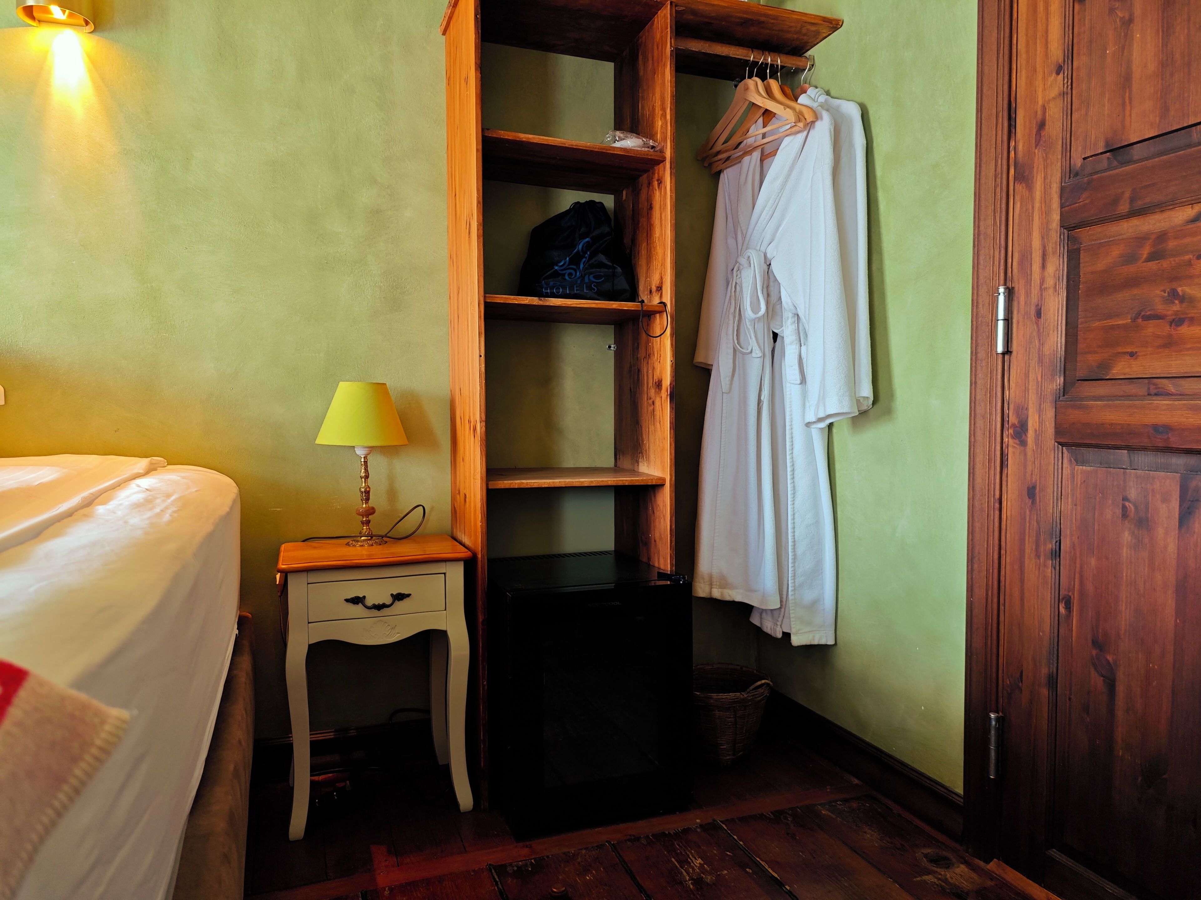 Una habitación con una cama, una mesita de noche de madera con lámpara, un armario con ropa colgada y una pequeña cesta.