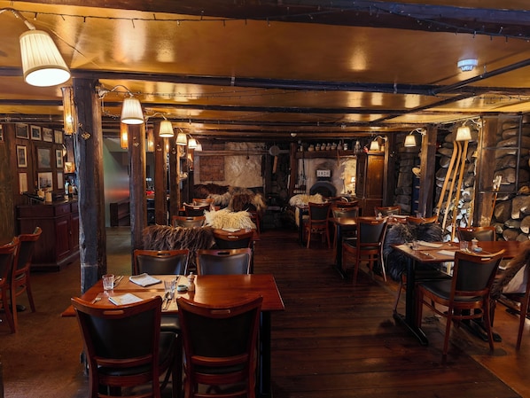 A restaurant interior with wooden tables and chairs, hanging lights, and a stone wall.