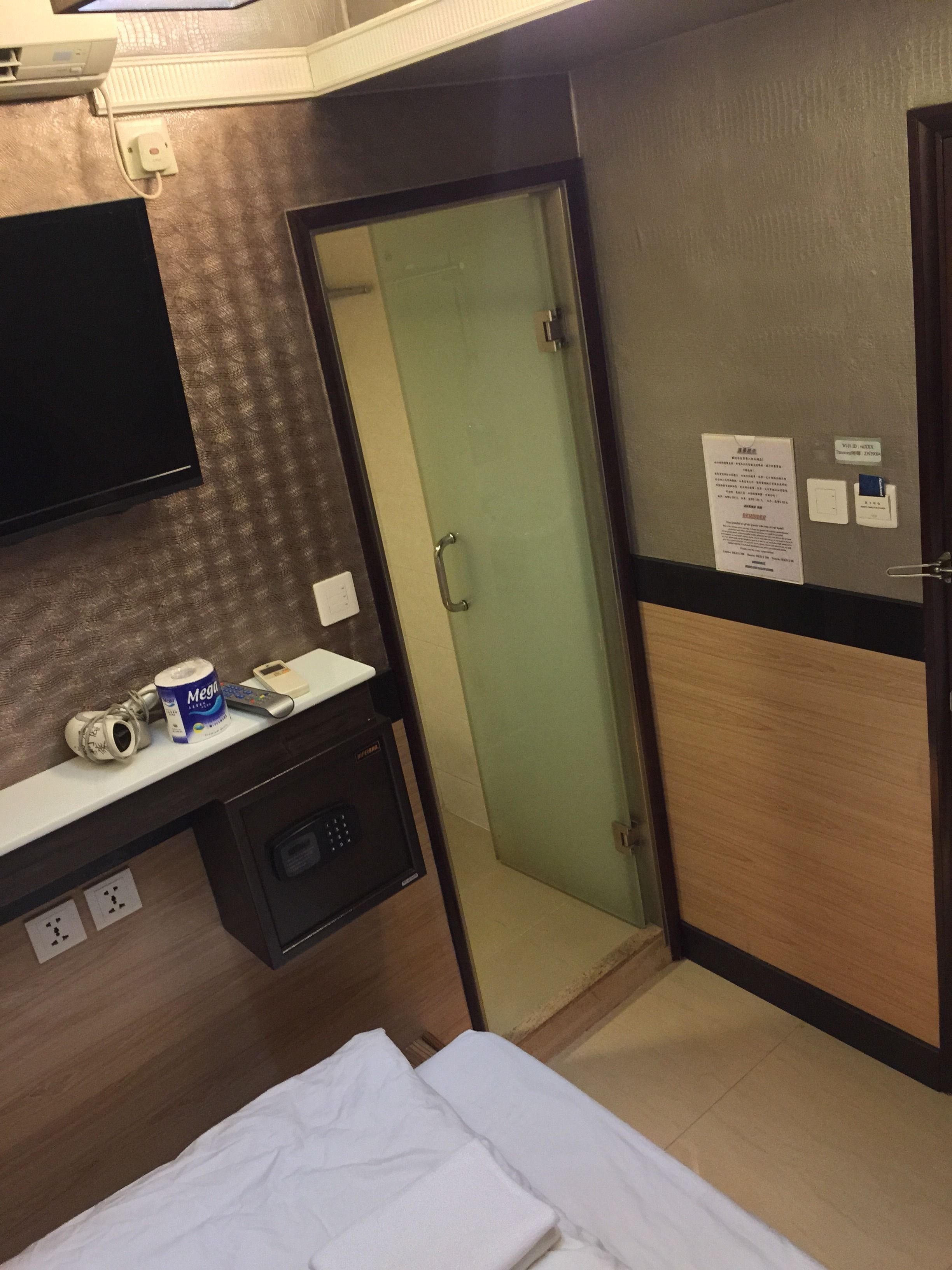 A hotel room with a bed, a desk with a computer, a small cabinet, a wall-mounted TV, and a door leading to another room.