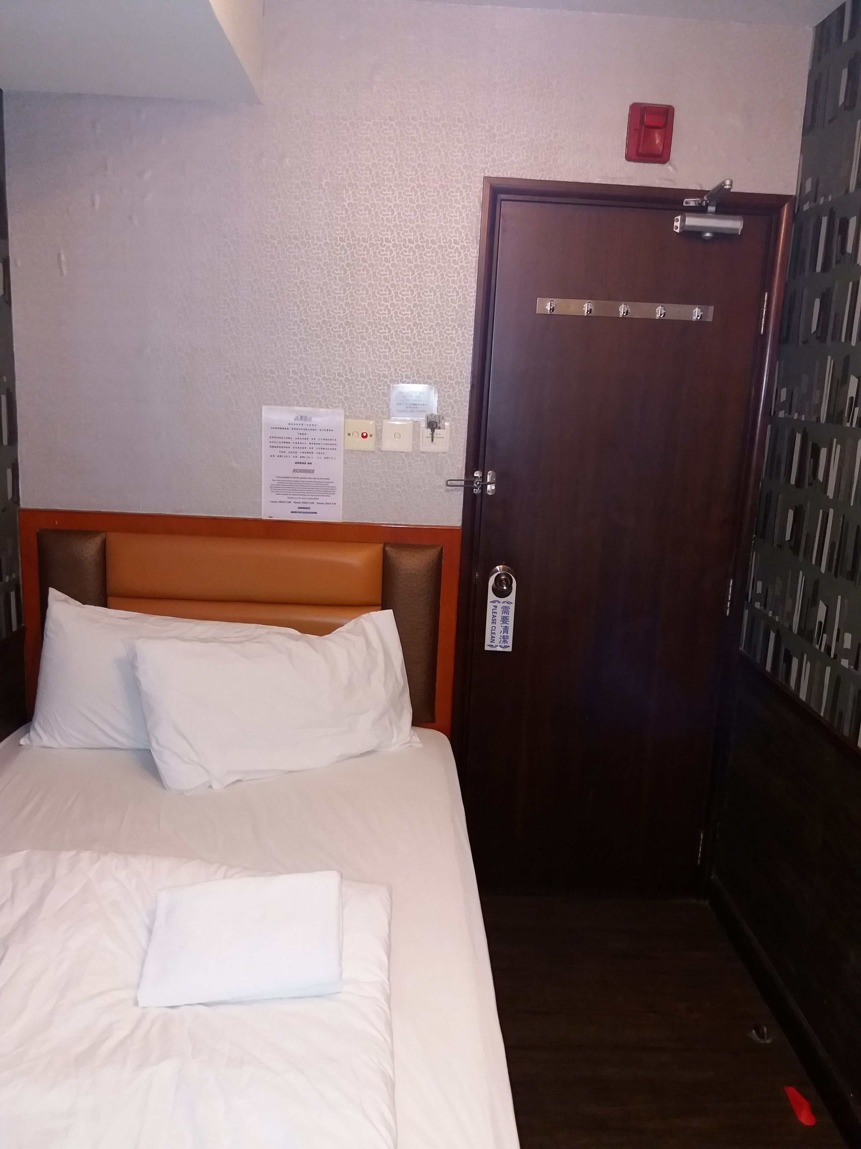 A hotel room with a bed, a wooden headboard, a door with a handle, and a wall-mounted light switch.