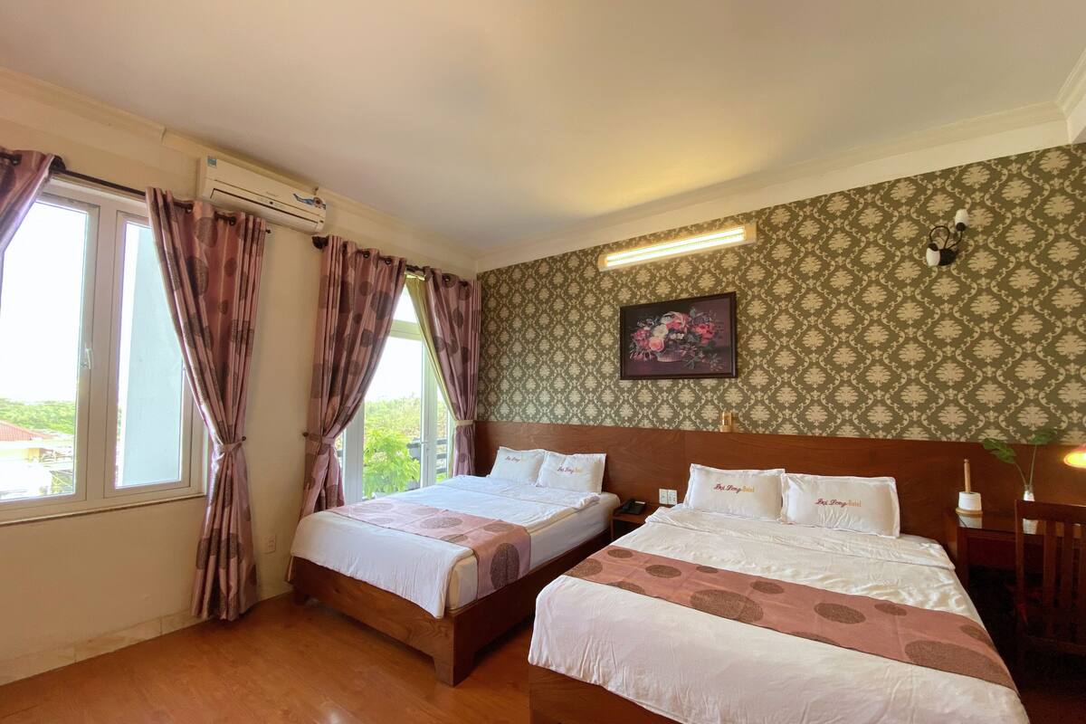 A hotel room with two beds, a window with curtains, an air conditioning unit, a wall-mounted light, and a floral painting.
