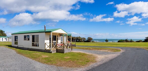 A small, single-story mobile home with a green roof and a porch, situated on a gravel driveway with a grassy area and a view of the ocean. - Discovery Parks - Devonport (East Devonport)