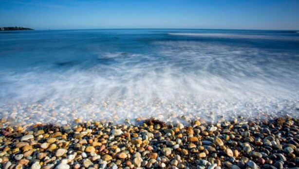 Une plage de galets avec une eau claire et une côte visible au loin.