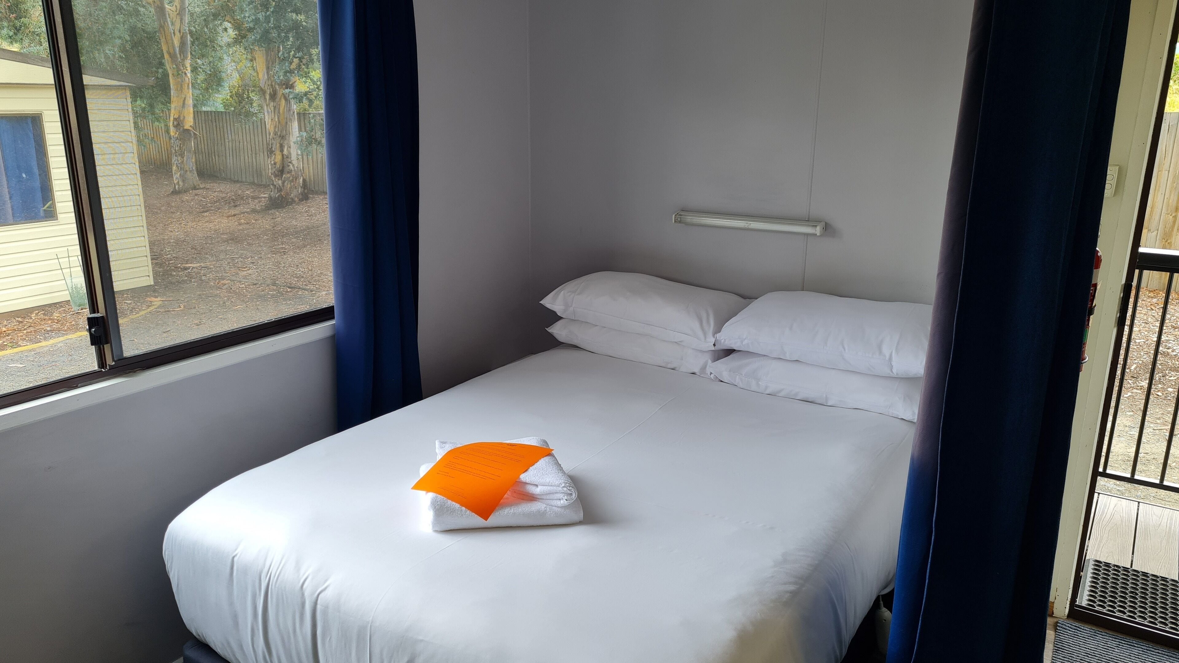 Un lit simple avec une literie blanche, un rideau bleu et une fenêtre donnant sur des arbres et une clôture.