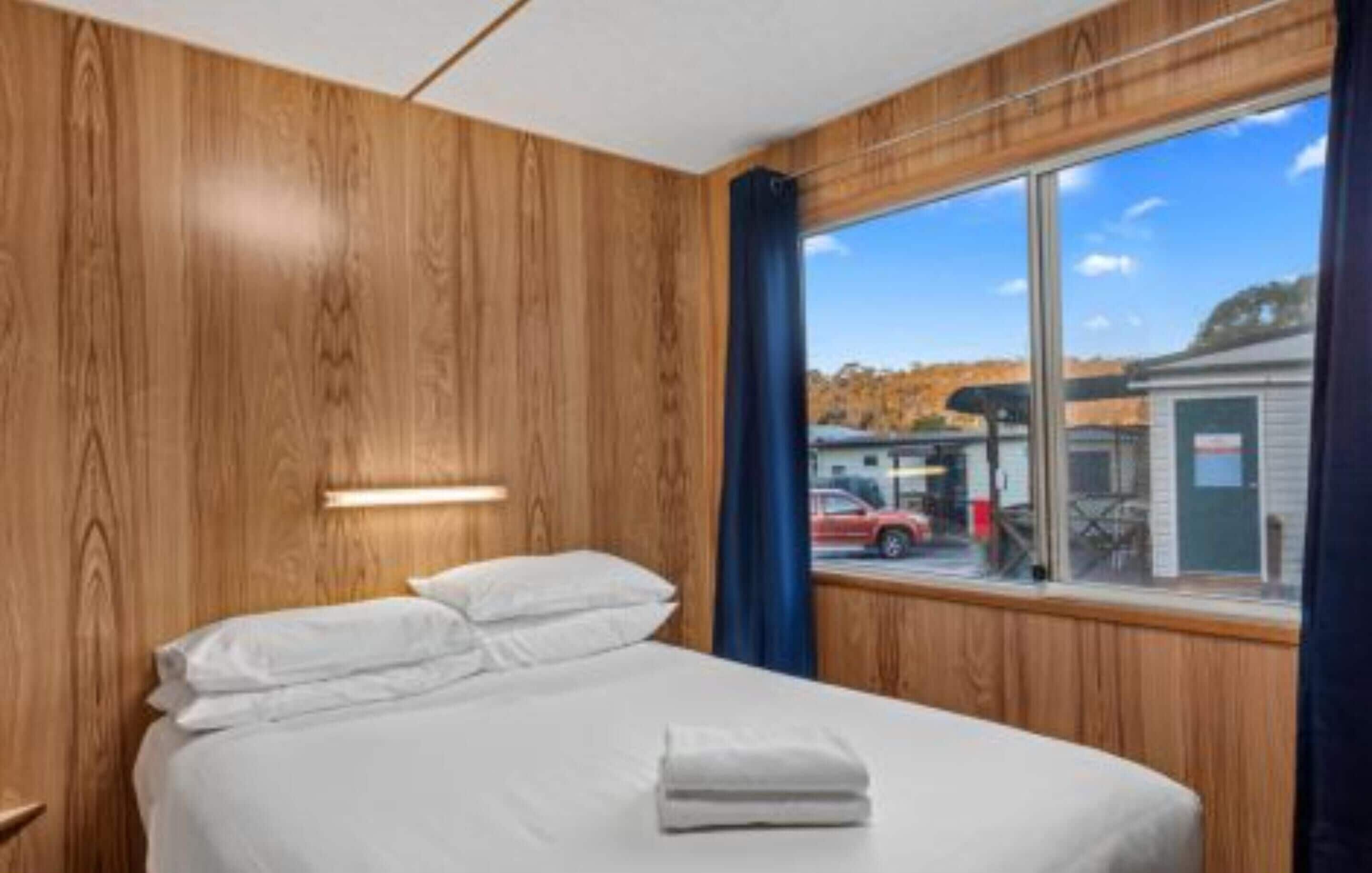 A hotel room with a wooden wall, a bed with white linens, and a large window offering a view of the outside.
