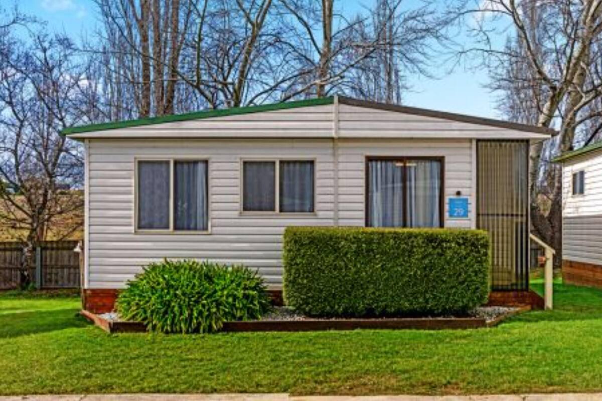 A single-story mobile home with a green roof, a small front yard with a hedge, and a blue handicap parking sign.