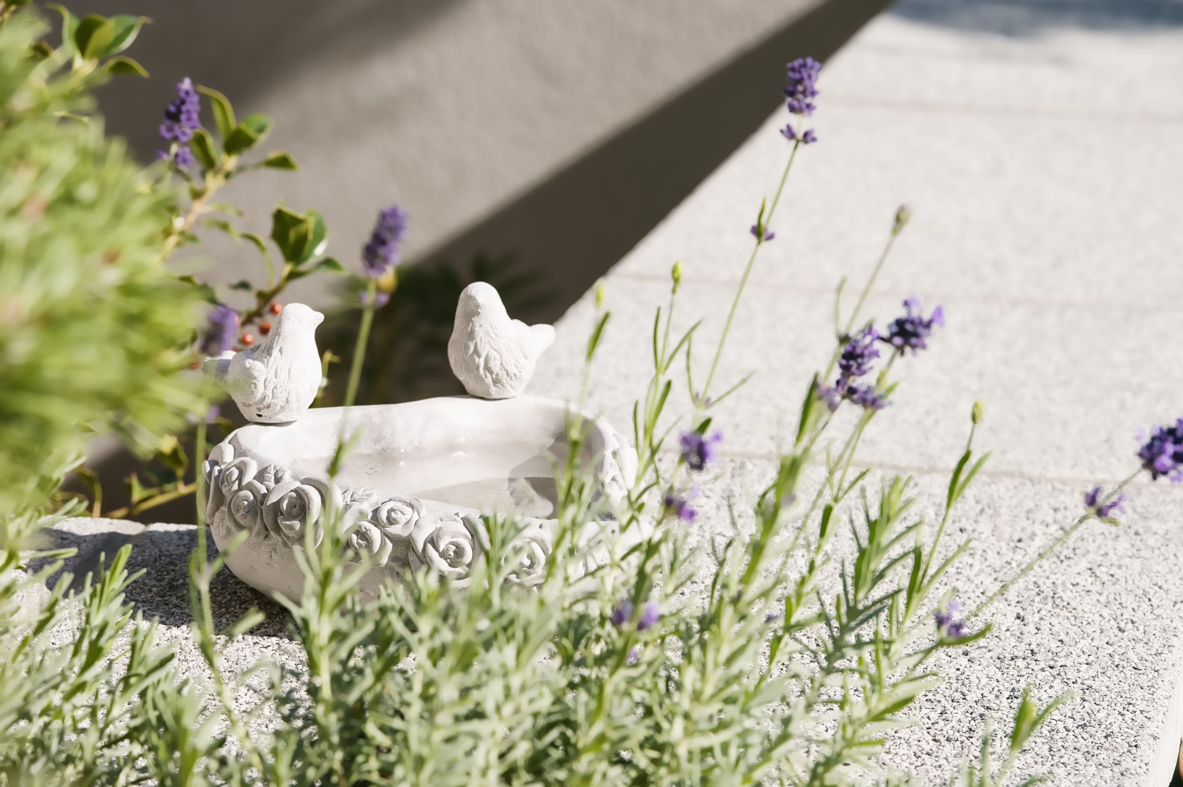 En hvid fuglebad i form af en fugl med detaljerede udskæringer, omgivet af grønt løv og lilla blomster.