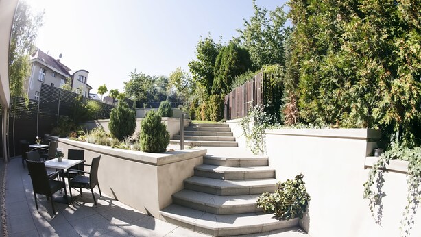 Un patio moderno al aire libre con zona de estar, una escalera que sube a un nivel superior y diversas plantas y árboles.