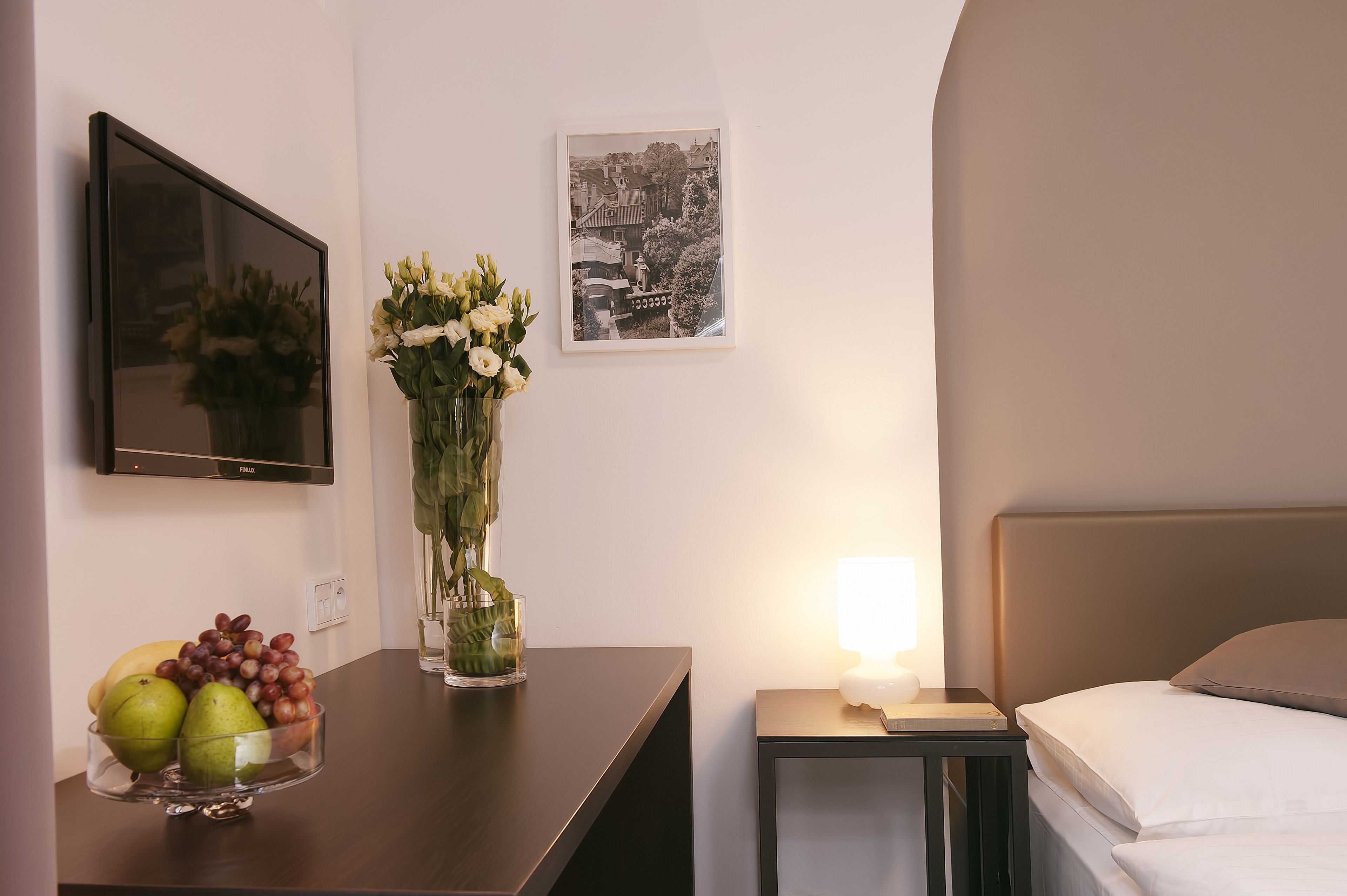 A hotel room with a bed, a bedside table with a lamp, a TV mounted on the wall, a framed picture, and a fruit bowl on the table.