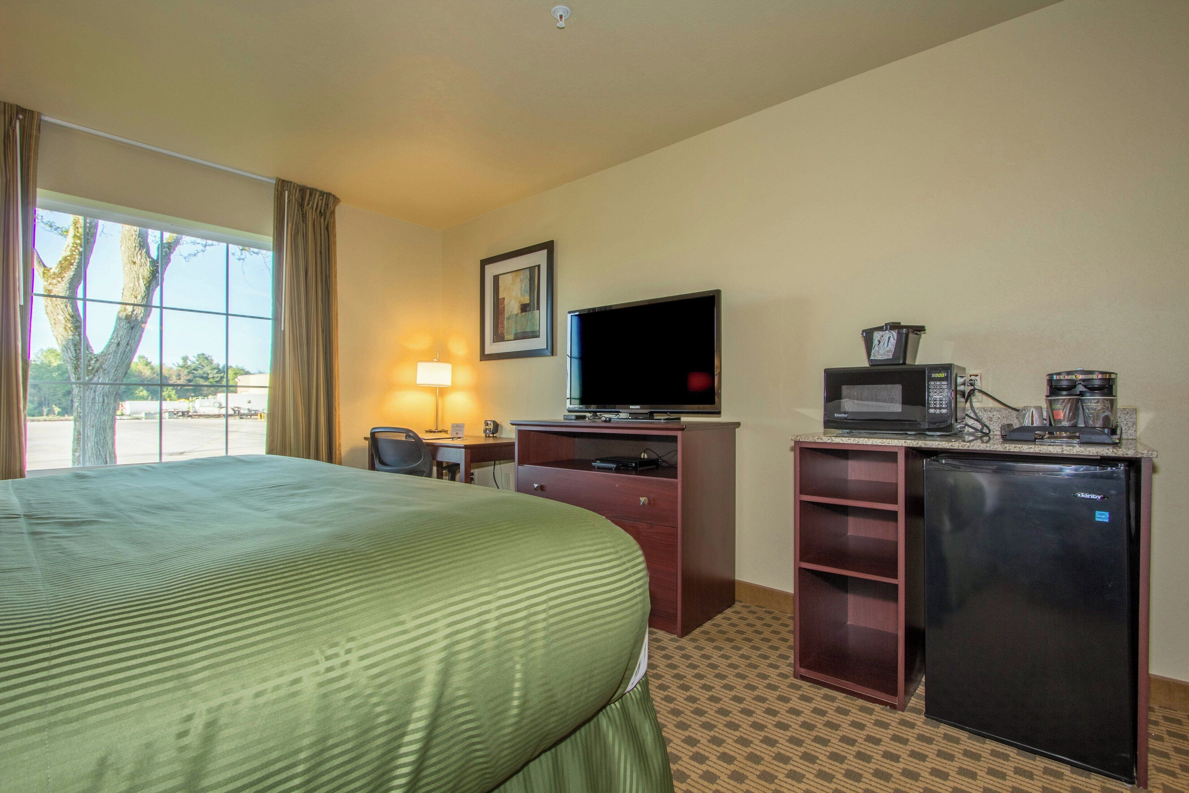 Una habitación de hotel con cama, escritorio con televisor, refrigerador y ventana con cortinas.