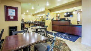 A hotel lobby with a reception desk, dining area with tables and chairs, a trash bin, and a framed certificate on the wall.