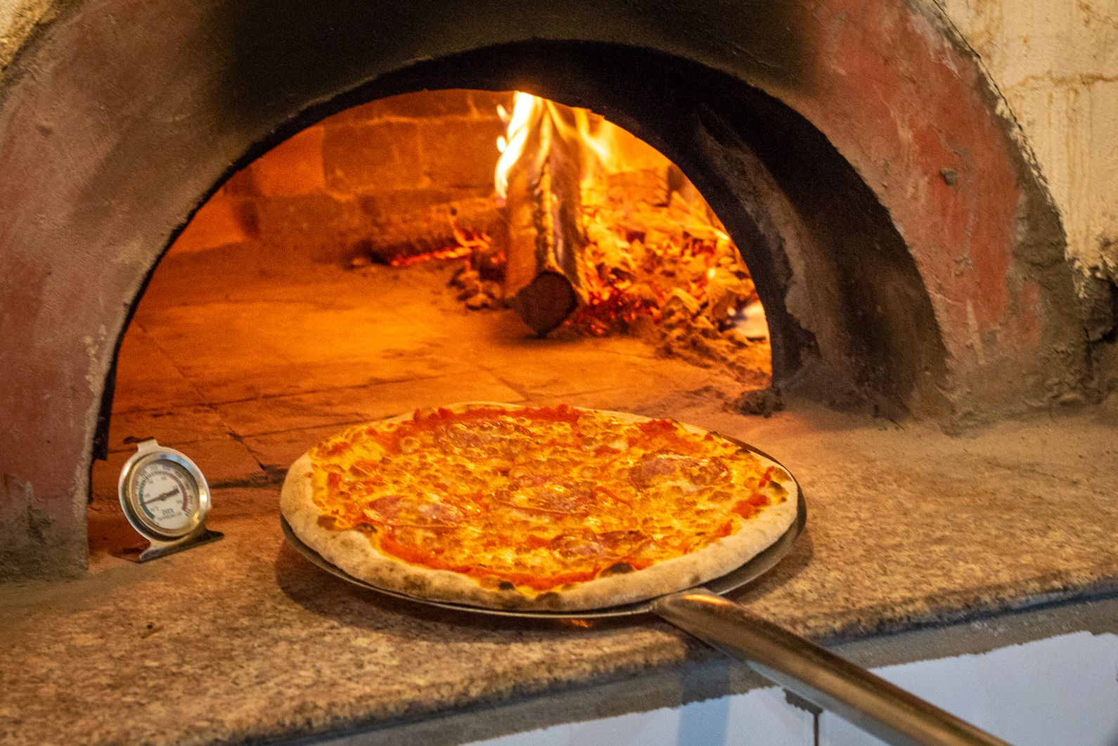 A pizza in a brick oven with a thermometer and a pizza peel.