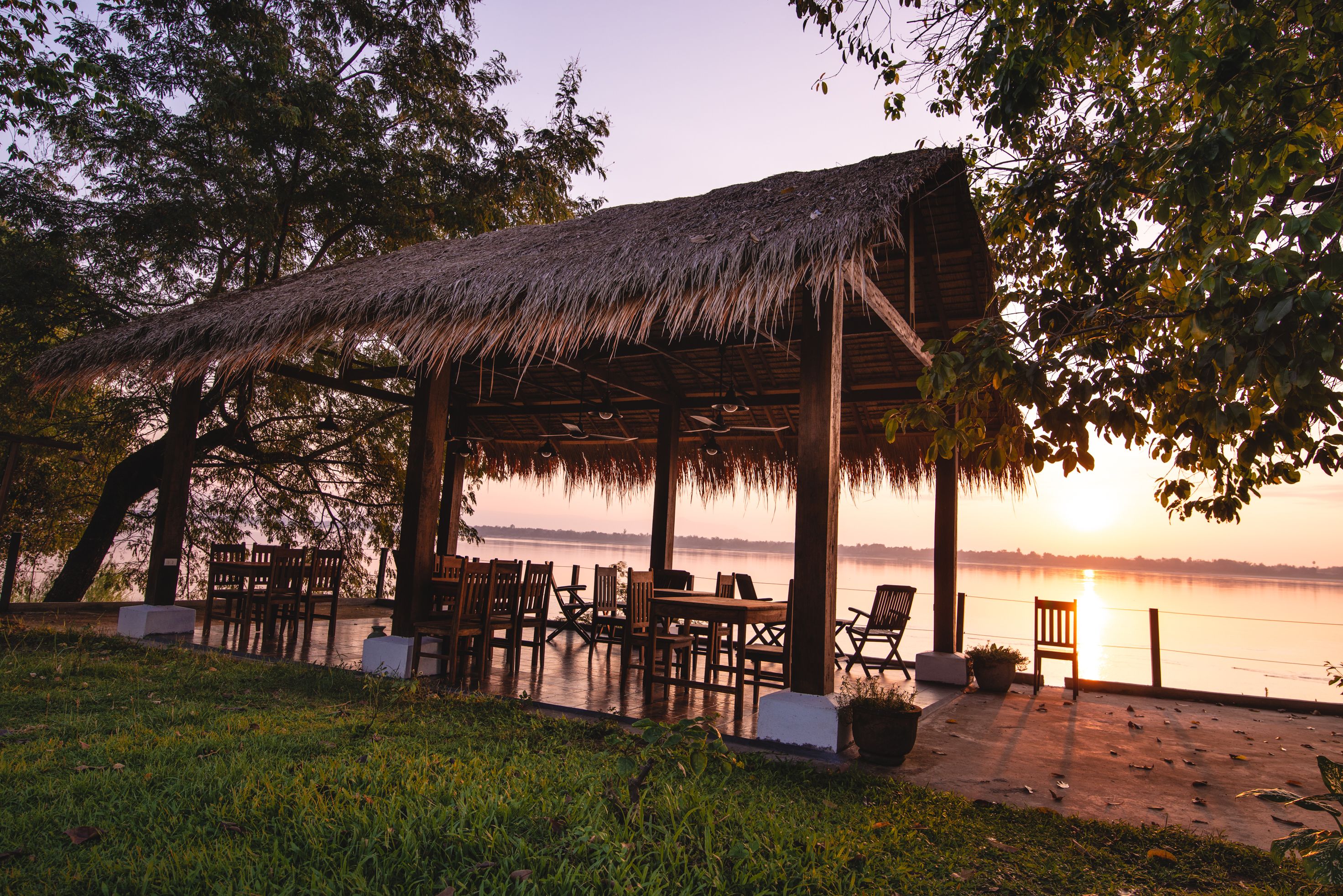 A thatched-roof pavilion with outdoor seating by a lake at sunset.