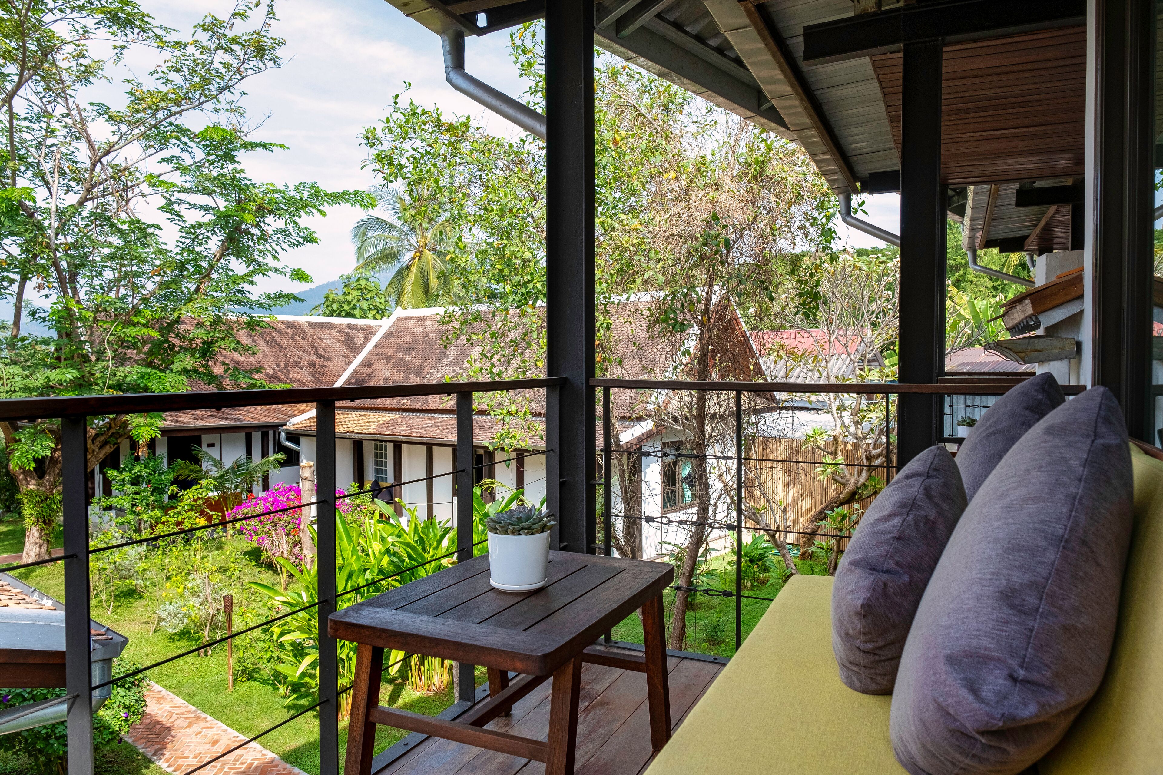 A balcony with a wooden table and cushions, overlooking a lush garden and traditional buildings.