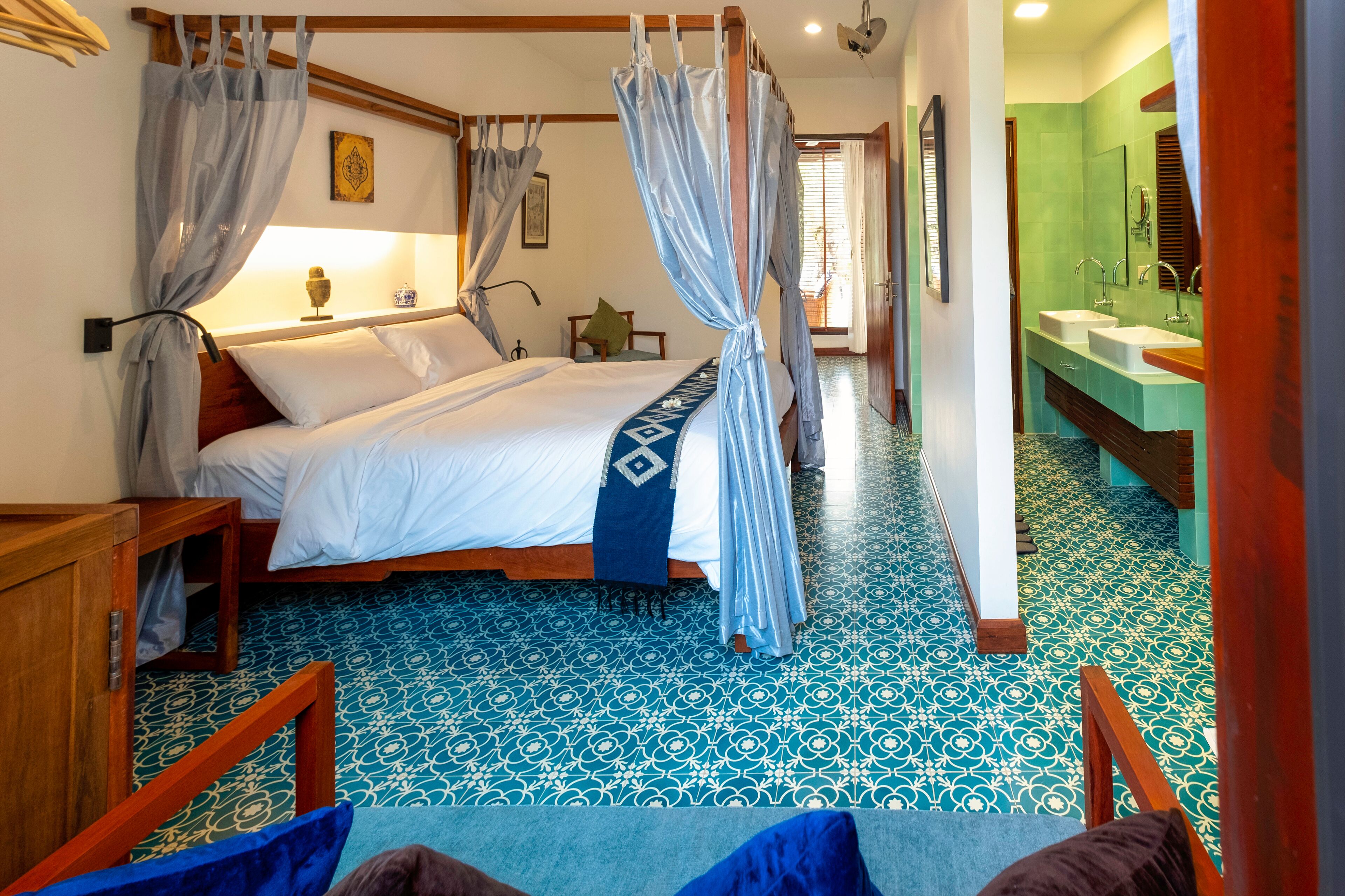 A room with a bed, a wooden nightstand, a blue and white patterned floor, and a bathroom with a green tiled wall and wooden counter.