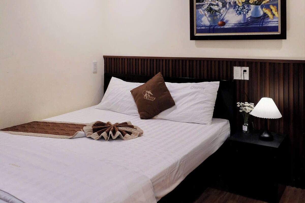 Ein ordentlich bezogenes Bett mit einer braunen Tagesdecke, ein gerahmtes Blumenbild an der Wand und ein kleiner Tisch mit einer Lampe und Blumen.