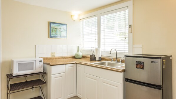 A compact kitchen with white cabinets, a stainless steel refrigerator, a microwave, and a window with blinds.
