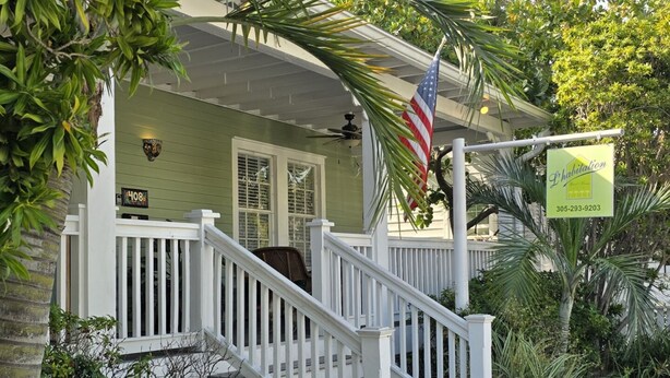 A green house with a white porch, a flag, and a sign advertising a property for sale. - L'Habitation Guest House - Adults Only (Key West)