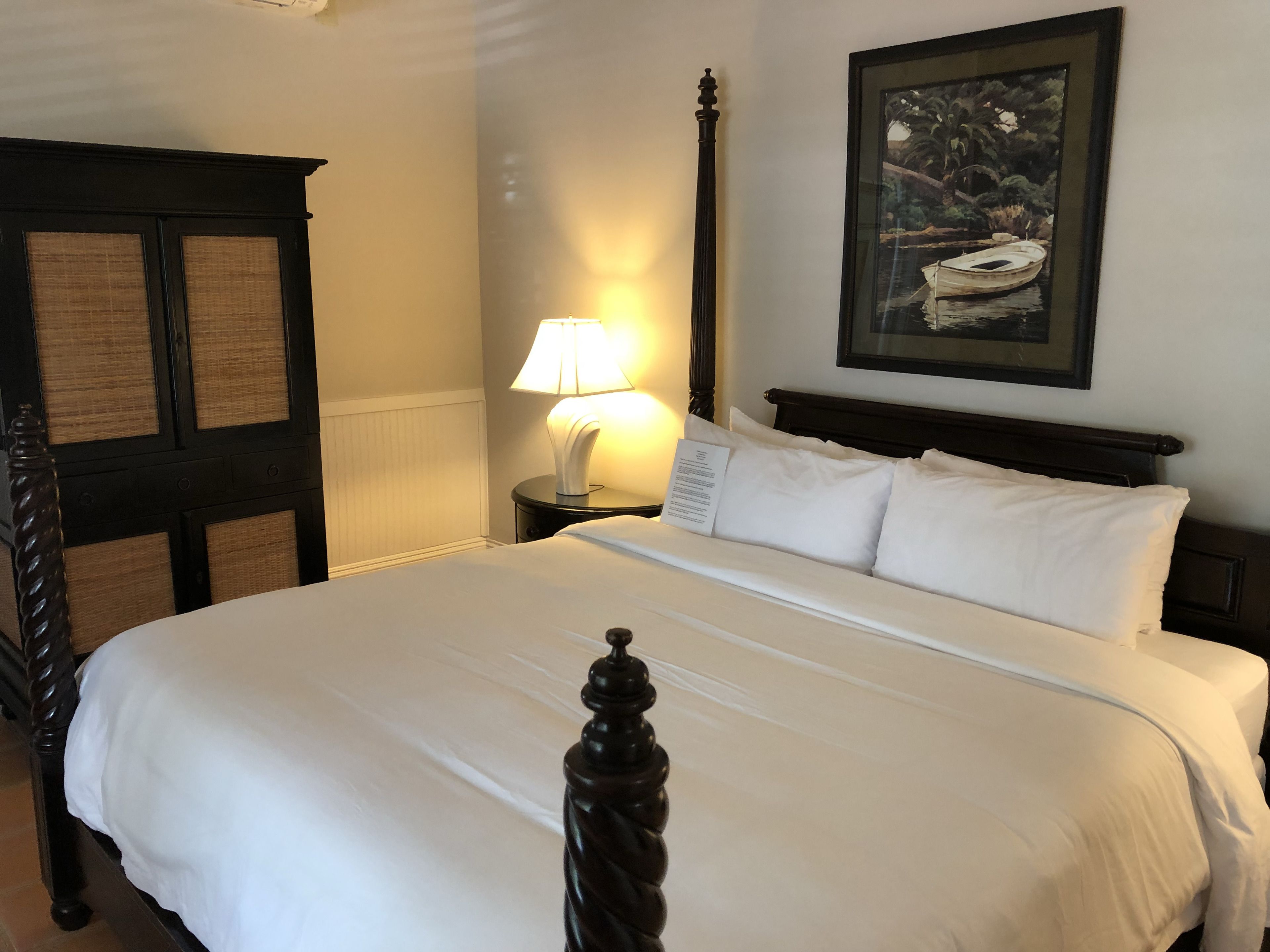 A neatly made bed with white linens, a dark wooden headboard, a nightstand with a lamp, and a framed picture on the wall.