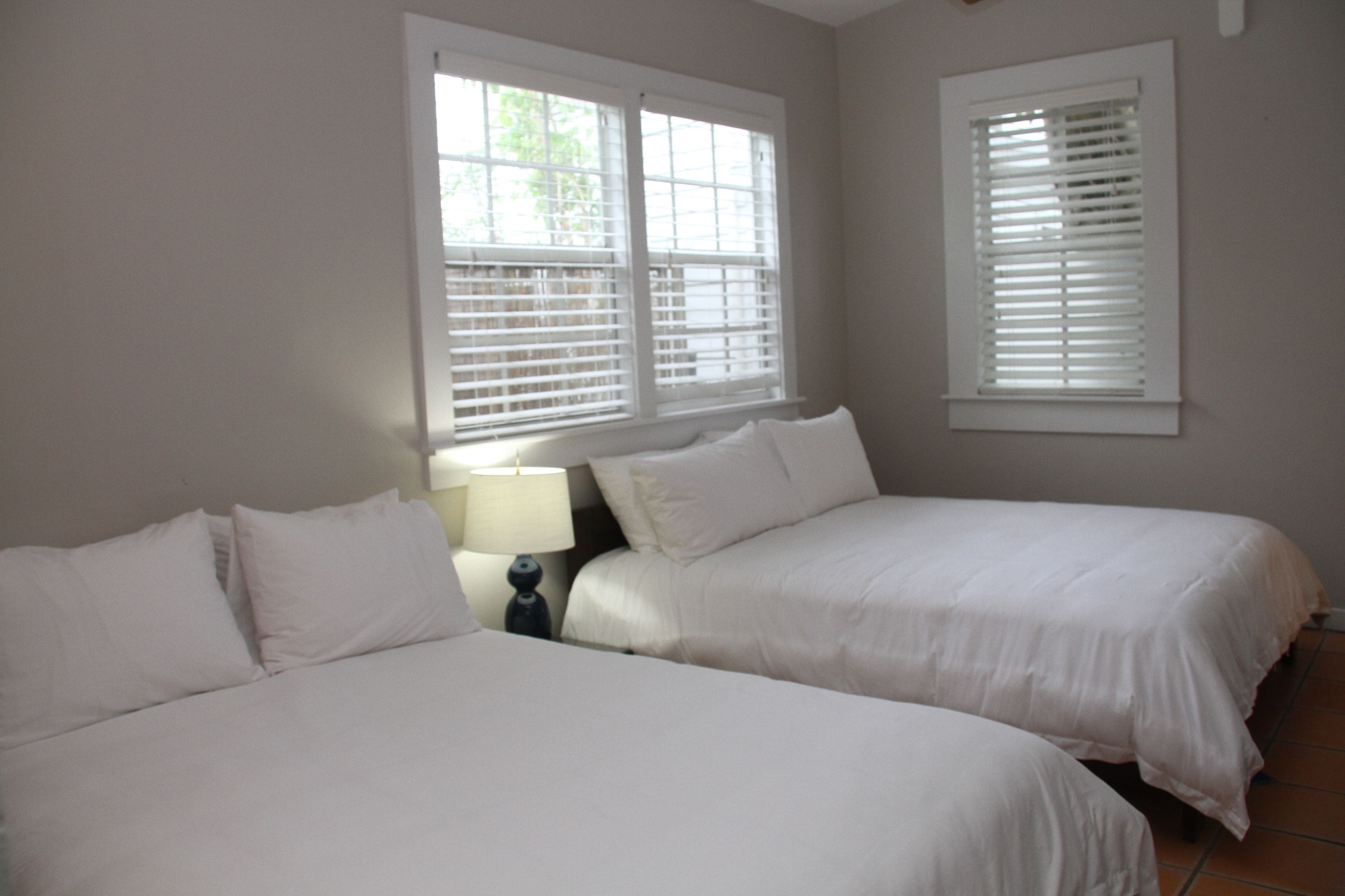 A bedroom with two beds, white bedding, a nightstand with a lamp, and windows with blinds.