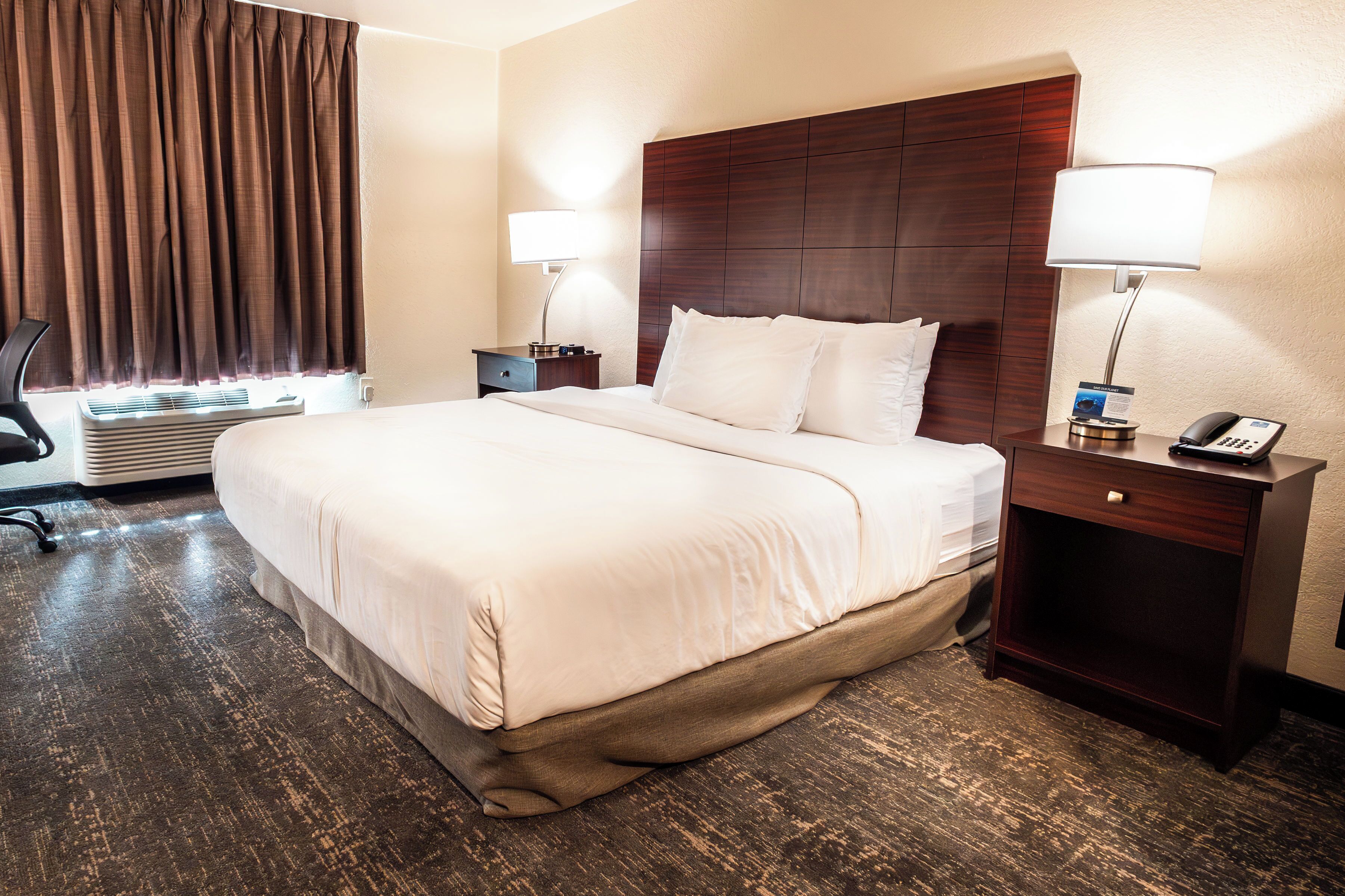 A hotel room with a large bed, two bedside tables with lamps, a desk with a chair, and a window with curtains.