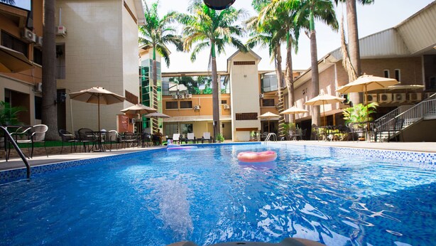 Una piscina con un flotador rosa, rodeada de sillas de descanso y sombrillas, con palmeras y un edificio de varios pisos al fondo.