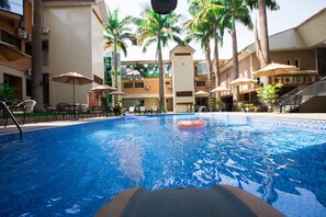 Une piscine avec un flotteur rose, entourée de chaises longues et de parasols, avec des palmiers et un immeuble à plusieurs étages en arrière-plan.