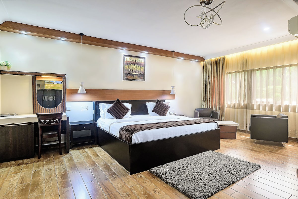 A modern hotel room with a wooden floor, a large bed with a headboard, a desk with a chair, a small table, and a large window with curtains.