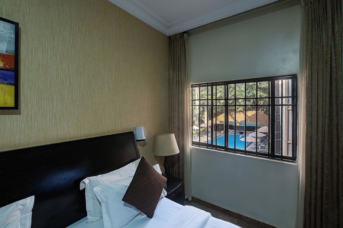 A hotel room with a bed, a nightstand with a lamp, a window with a view of a pool and outdoor furniture.